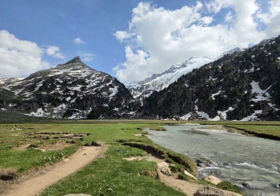 Ruta de 5 días por Benasque. Un riachuelo cristalino fluye a través de la verde y llana pradera alpina de Aigualluts, con un sendero de tierra en primer plano y majestuosas montañas nevadas al fondo bajo un cielo azul con nubes blancas.