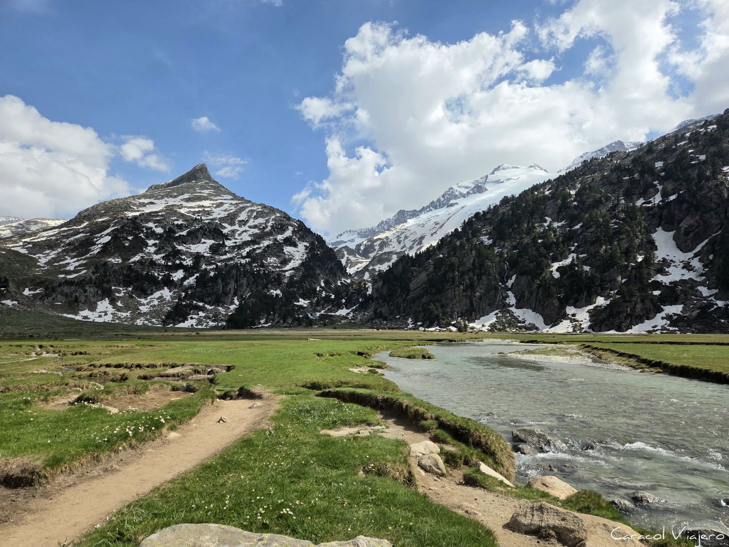Ruta de 5 días por Benasque. Un riachuelo cristalino fluye a través de la verde y llana pradera alpina de Aigualluts, con un sendero de tierra en primer plano y majestuosas montañas nevadas al fondo bajo un cielo azul con nubes blancas.
